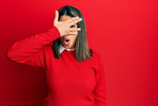 Young latin woman wearing casual clothes peeking in shock covering face and eyes with hand, looking through fingers with embarrassed expression.  - Photo, Image