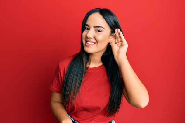 A fiatal, gyönyörű spanyol lány alkalmi piros pólóban mosolyog, fültől fülig hallgatva a pletykákat vagy pletykákat. süketség fogalma.  - Fotó, kép