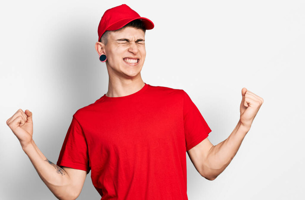 Young caucasian boy with ears dilation wearing delivery uniform and cap very happy and excited doing winner gesture with arms raised, smiling and screaming for success. celebration concept.  - Photo, Image