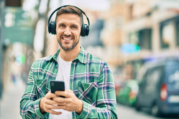 młody biały mężczyzna uśmiechnięty szczęśliwy za pomocą smartfona i słuchawek w mieście. - Zdjęcie, obraz