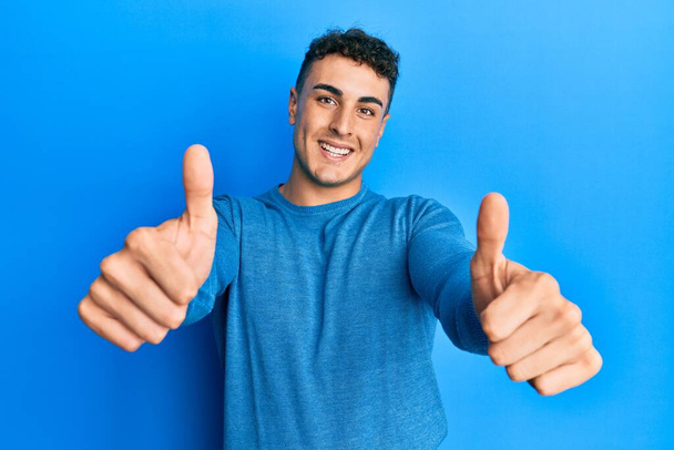 Hispanischer junger Mann in lässigem Winterpullover, der eine positive Geste mit der Hand zustimmt, Daumen hoch lächelnd und glücklich über den Erfolg. Siegergeste.  - Foto, Bild