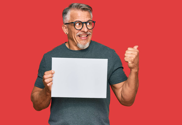 Middle age grey-haired man holding blank empty banner pointing thumb up to the side smiling happy with open mouth  - Foto, Bild