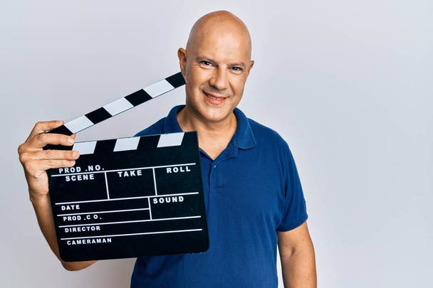 Hombre calvo de mediana edad sosteniendo el tablero de vídeo de la película que mira positivo y feliz de pie y sonriendo con una sonrisa confiada que muestra los dientes  - Foto, imagen