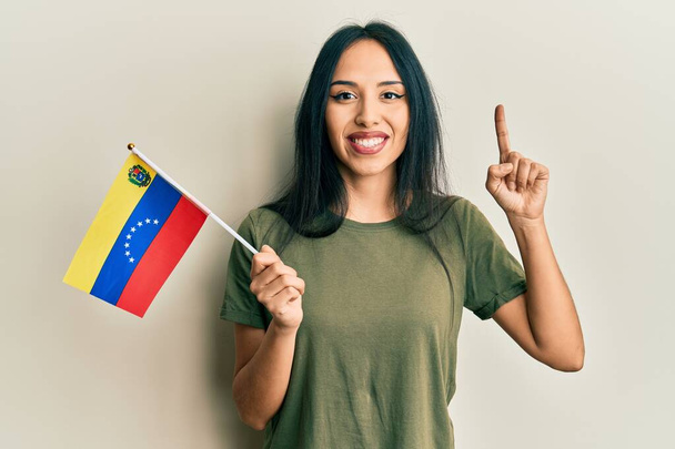 Νεαρή Ισπανίδα που κρατά τη σημαία της Βενεζουέλας χαμογελώντας με μια ιδέα ή ερώτηση δείχνοντας το δάχτυλο με χαρούμενο πρόσωπο, νούμερο ένα  - Φωτογραφία, εικόνα