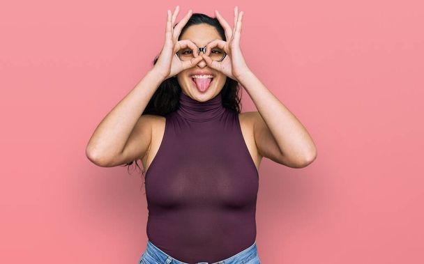 Brunette young woman wearing casual clothes and glasses doing ok gesture like binoculars sticking tongue out, eyes looking through fingers. crazy expression.  - Фото, зображення