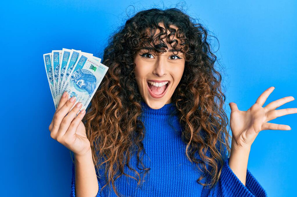 Young hispanic girl holding 50 polish zloty banknotes celebrating achievement with happy smile and winner expression with raised hand  - Foto, afbeelding