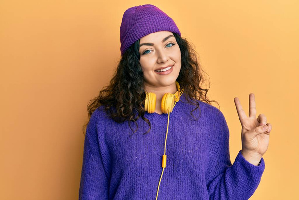 Young brunette woman with curly hair listening to music using headphones showing and pointing up with fingers number two while smiling confident and happy.  - Fotoğraf, Görsel