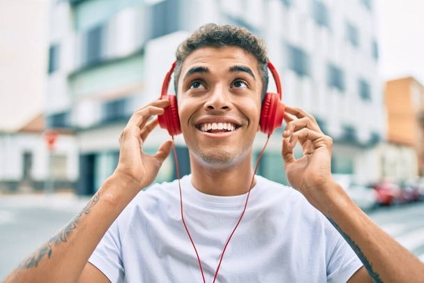 Jovem latino sorrindo feliz usando fones de ouvido andando na cidade. - Foto, Imagem