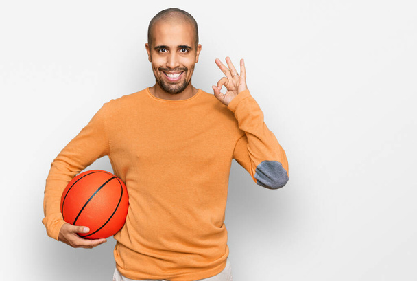 Latynoski dorosły mężczyzna trzyma piłkę do koszykówki robi ok znak palcami, uśmiechnięty przyjazny gest doskonały symbol  - Zdjęcie, obraz