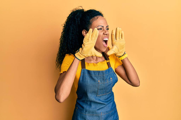 Medioevo donna afroamericana indossando grembiule professionale urlando arrabbiato ad alta voce con le mani sopra la bocca  - Foto, immagini