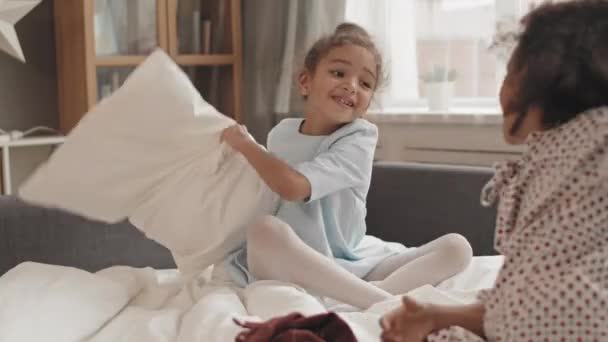 Повне надмірне плече радісної змішаної раси маленька дівчинка, яка має подушку боротися з невпізнаваним другом на ліжку в кімнаті
 - Кадри, відео