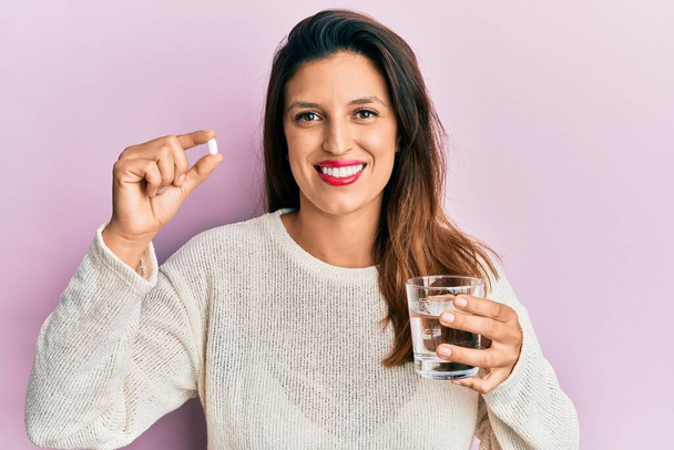 Красива іспаномовна жінка тримає таблетку і склянку води посміхається з щасливою і холодною посмішкою на обличчі. показ зубів
.  - Фото, зображення