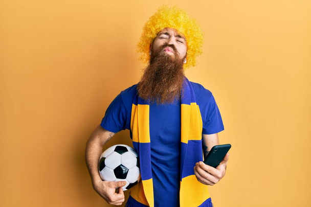 Pelirrojo hombre con barba larga hooligan fútbol animando juego sosteniendo smartphone mirando a la cámara soplando un beso siendo encantador y sexy. expresión de amor.  - Foto, Imagen