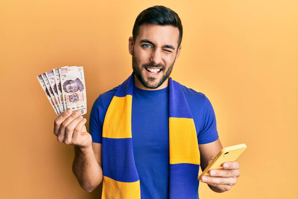 Młody Hiszpan kibic kibicujący piłce nożnej za pomocą smartfona trzymającego meksykańskie peso mrugające banknoty patrzące w kamerę z seksowną ekspresją, wesołą i szczęśliwą twarzą.  - Zdjęcie, obraz