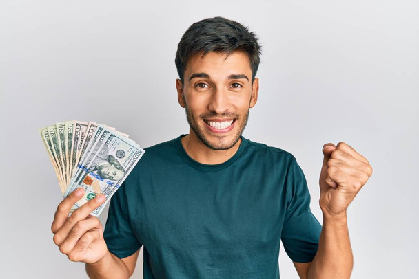 Nuori komea mies pitää dollareita huutaen ylpeä, juhlii voittoa ja menestystä hyvin innoissaan kohotettu käsi  - Valokuva, kuva