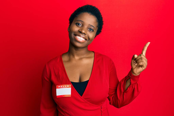 Νεαρή Αφρικάνα με κοντά μαλλιά φορώντας καλημέρα το όνομά μου είναι αυτοκόλλητη ταυτότητα χαμογελώντας χαρούμενη δείχνοντας με το χέρι και το δάχτυλο στο πλάι.  - Φωτογραφία, εικόνα