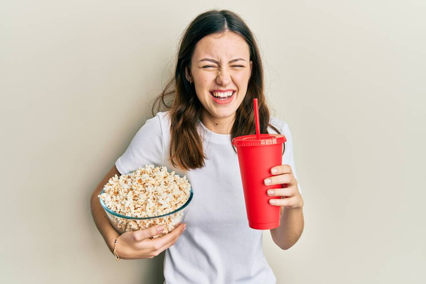 Young brunette woman eating popcorn and drinking soda smiling and laughing hard out loud because funny crazy joke.  - Photo, Image