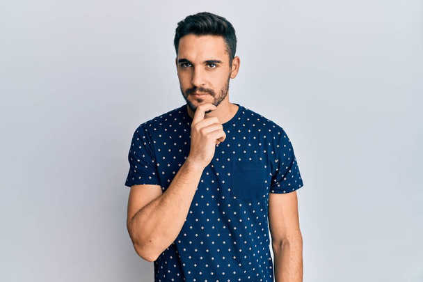 Fiatal spanyol férfi alkalmi ruhát visel gondolkodás koncentrált kétség ujjal az állán, és felnézett csodálkozva  - Fotó, kép