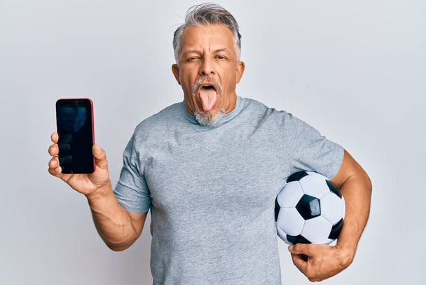 Grauhaariger Mann mittleren Alters hält Fußballball in die Höhe und zeigt Smartphone, das mit lustigem Gesichtsausdruck die Zunge herausstreckt.  - Foto, Bild