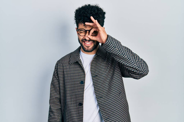 Young arab man with beard wearing glasses doing ok gesture with hand smiling, eye looking through fingers with happy face.  - Valokuva, kuva