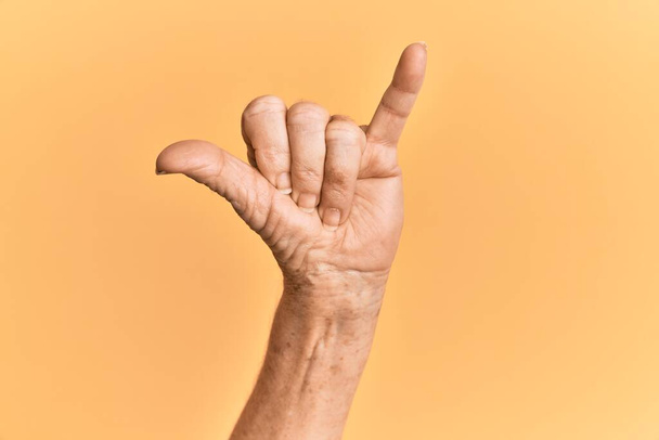 Starší kavkazská ruka nad žlutým izolovaným pozadím gestikulující havajské gesto na pozdrav, telefon a komunikační symbol  - Fotografie, Obrázek