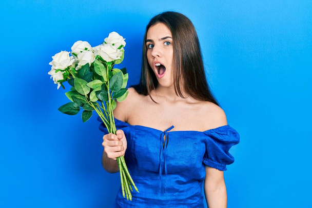 Young brunette teenager holding bouquet of white flowers scared and amazed with open mouth for surprise, disbelief face  - Foto, imagen