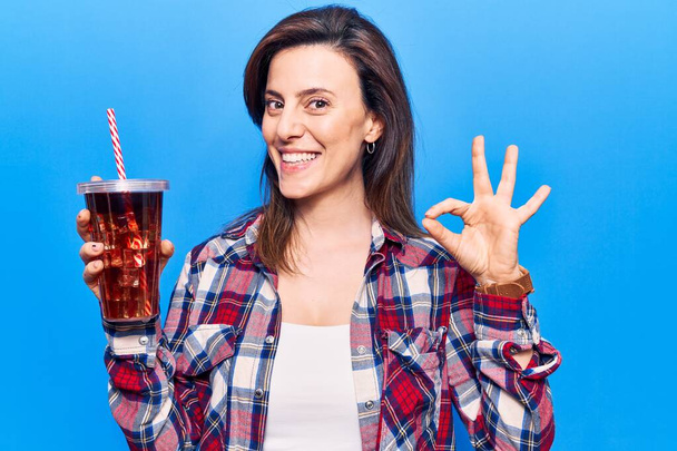 Giovane bella donna bere bicchiere di cola bevanda facendo segno ok con le dita, sorridente gesto amichevole eccellente simbolo  - Foto, immagini