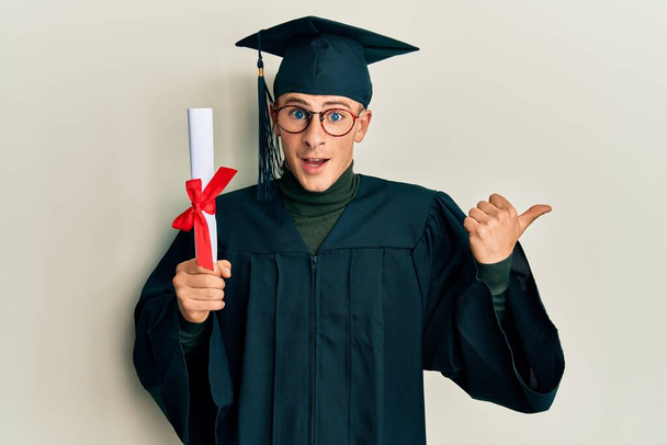 Young caucasian man wearing graduation cap and ceremony robe holding diploma pointing thumb up to the side smiling happy with open mouth  - Foto, Imagen