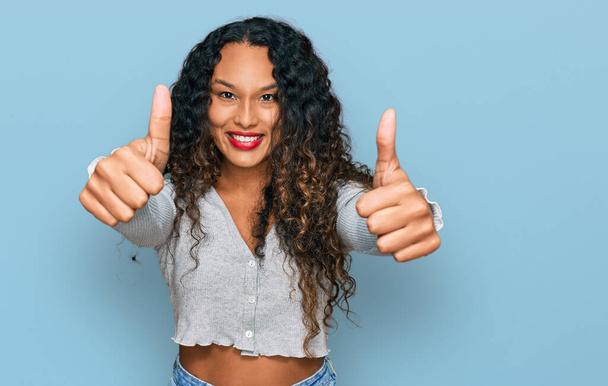 Young hispanic woman with curly hair wearing casual clothes approving doing positive gesture with hand, thumbs up smiling and happy for success. winner gesture.  - Photo, Image