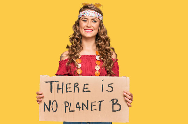 Young blonde girl wearing hippie style holding there is no planet b banner looking positive and happy standing and smiling with a confident smile showing teeth  - 写真・画像