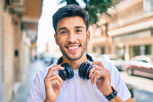 Jovem latino sorrindo feliz ouvindo música usando fones de ouvido na cidade. - Foto, Imagem