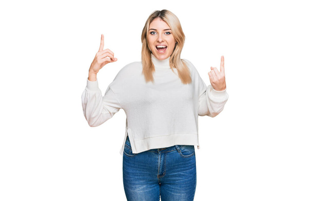 Jonge blanke vrouw draagt casual winter trui glimlachend verbaasd en verrast en wijzen omhoog met vingers en opgeheven armen.  - Foto, afbeelding