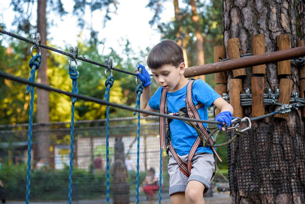 Young cute child boy in summer clothing, safety harness and helmet attached with carbine to cable moves slowly along rope way on green trees sunny bokeh background. Sport, game, leisure concept. - Photo, Image