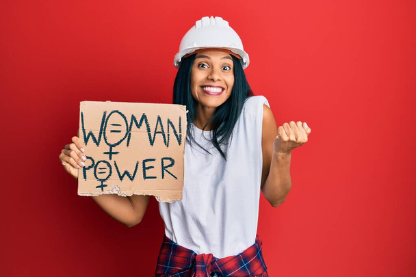 Young african american woman wearing architect hardhat holding woman power banner screaming proud, celebrating victory and success very excited with raised arm  - Photo, Image