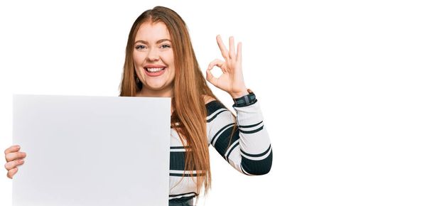 Młoda piękna ruda kobieta trzyma pusty sztandar robi ok znak palcami, uśmiechając przyjazny gest doskonały symbol  - Zdjęcie, obraz