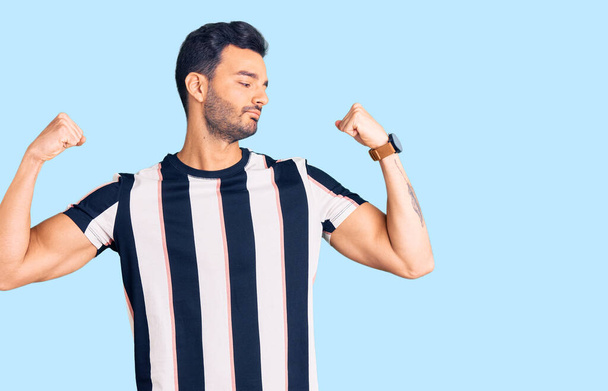 Ein junger gutaussehender hispanischer Mann in gestreiftem T-Shirt, der stolz lächelnd die Armmuskeln zeigt. Fitnesskonzept.  - Foto, Bild