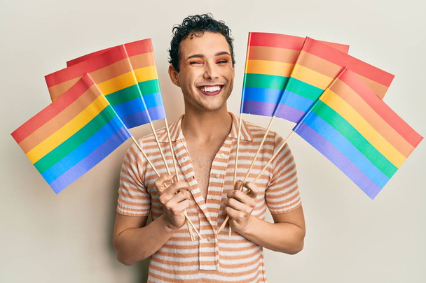 Handsome man wearing make up holding rainbow lgbtq flags winking looking at the camera with sexy expression, cheerful and happy face.  - Foto, Imagen