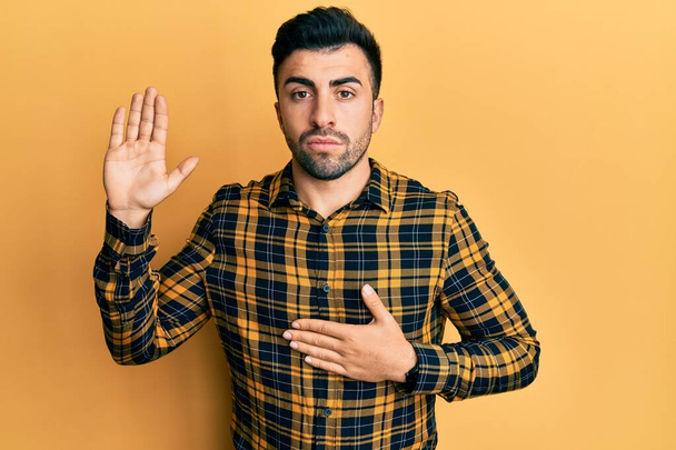 Joven hombre hispano usando ropa casual jurando con la mano en el pecho y la palma abierta, haciendo un juramento de lealtad  - Foto, imagen