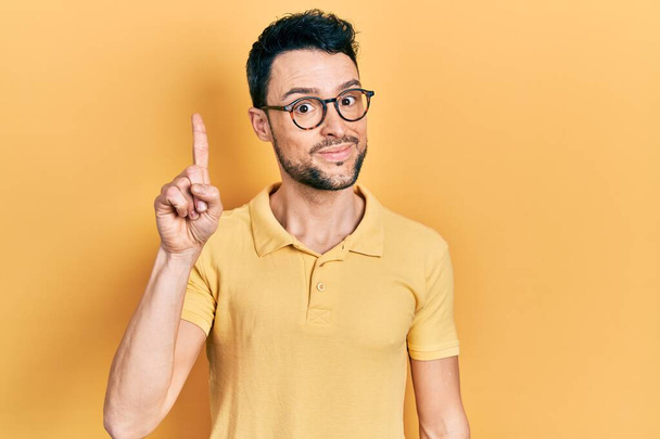 Νεαρός Ισπανός άνδρας που φοράει καθημερινά ρούχα και γυαλιά δείχνοντας και δείχνοντας προς τα πάνω με το δάχτυλο νούμερο ένα χαμογελώντας σίγουρος και χαρούμενος.  - Φωτογραφία, εικόνα