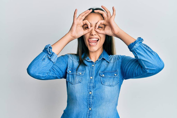 Young hispanic woman wearing casual denim jacket doing ok gesture like binoculars sticking tongue out, eyes looking through fingers. crazy expression.  - Foto, imagen