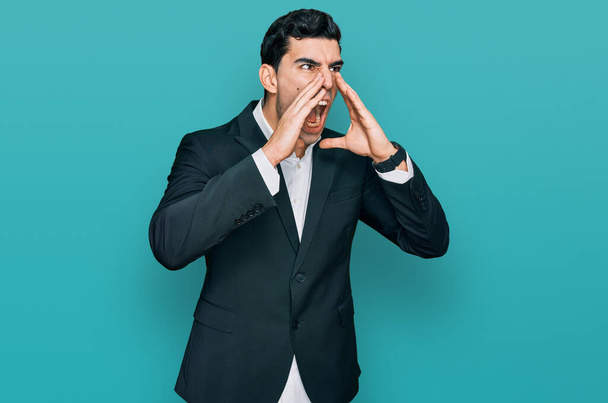 Schöner hispanischer Mann in Businesskleidung schreit wütend laut mit den Händen über dem Mund  - Foto, Bild