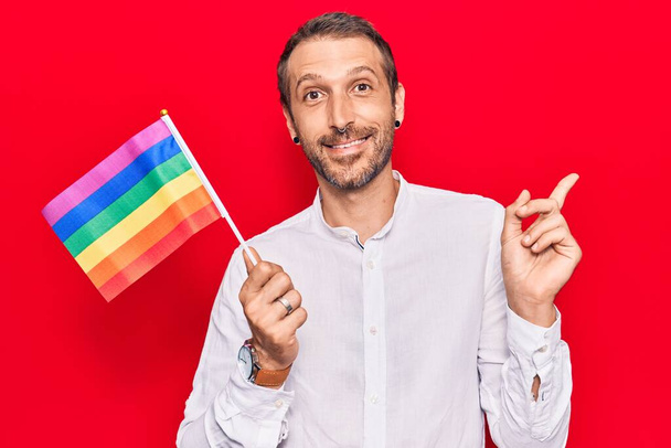 Jonge knappe man met regenboog lgbtq vlag lachend gelukkig wijzend met hand en vinger naar de zijkant  - Foto, afbeelding