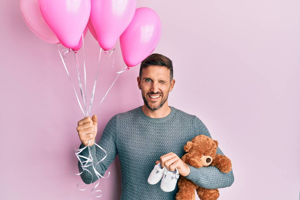 Handsome man with beard expecting a baby girl holding balloons, shoes and teddy bear winking looking at the camera with sexy expression, cheerful and happy face.  - Photo, image