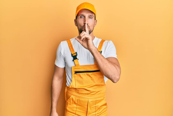 Young handsome man wearing handyman uniform over yellow background asking to be quiet with finger on lips. silence and secret concept.  - Fotoğraf, Görsel