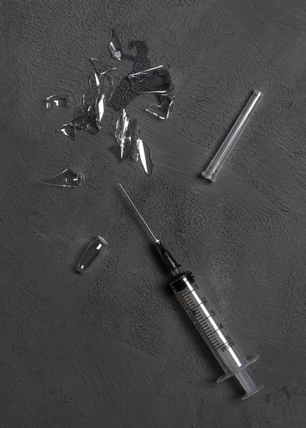 Použitá jednorázová injekční stříkačka a rozbitá skleněná ampulka s dávkou léku na šedém betonovém podkladu. - Fotografie, Obrázek
