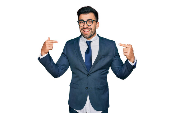 Hispanischer Mann mit Bart in Businessanzug und Krawatte sieht selbstbewusst aus mit einem Lächeln auf dem Gesicht, zeigt mit den Fingern stolz und glücklich.  - Foto, Bild