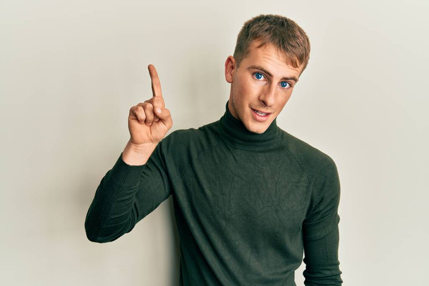 Νεαρός Καυκάσιος που φοράει καθημερινά ρούχα χαμογελώντας με μια ιδέα ή ερώτηση δείχνοντας το δάχτυλο του με χαρούμενο πρόσωπο, νούμερο ένα  - Φωτογραφία, εικόνα