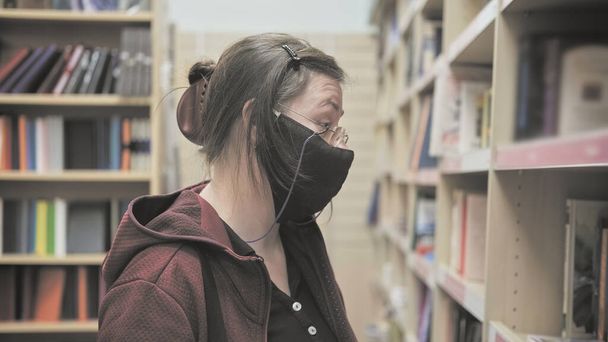 Nainen lasit ja naamio virusta vastaan katsoo kirjahylly varastossa ja yllättynyt - Valokuva, kuva