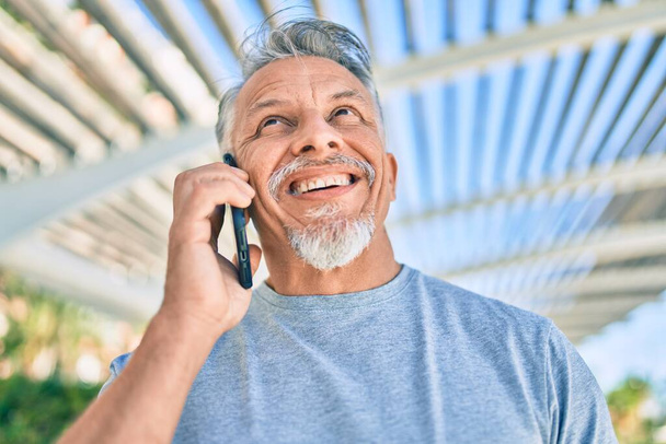 Homem hispânico de cabelos grisalhos de meia idade sorrindo feliz falando no smartphone no parque. - Foto, Imagem