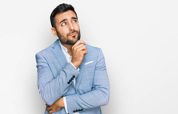 Junger hispanischer Mann in Businessjacke mit ernstem Gesicht, der mit der Hand am Kinn über Fragen nachdenkt, nachdenklich über verwirrende Ideen  - Foto, Bild
